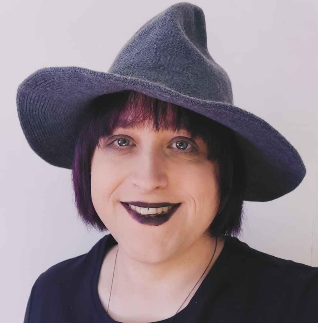 Lily smiling in a black outfit and gray hat against a plain background.